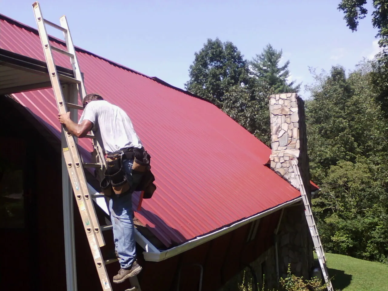 Roofer on a ladder beside a red metal roof and stone chimney — Soffit & Fascia Repair by Roofing Brothers in Estill Springs